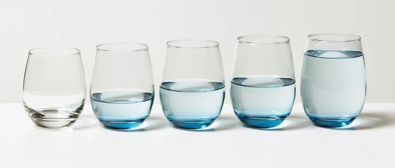 Six stemless drinking glasses in a row filled with increasing amounts of blue water on a white surface