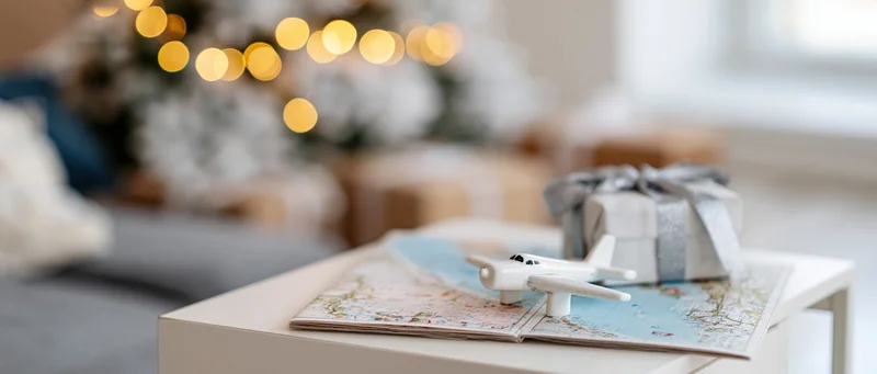 Small white toy airplane resting on open paper travel map on coffee table near christmas tree with wrapped gift and festive lights in background