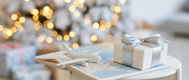 Small white toy airplane resting on open travel map with wrapped gift beside it on table near decorated christmas tree