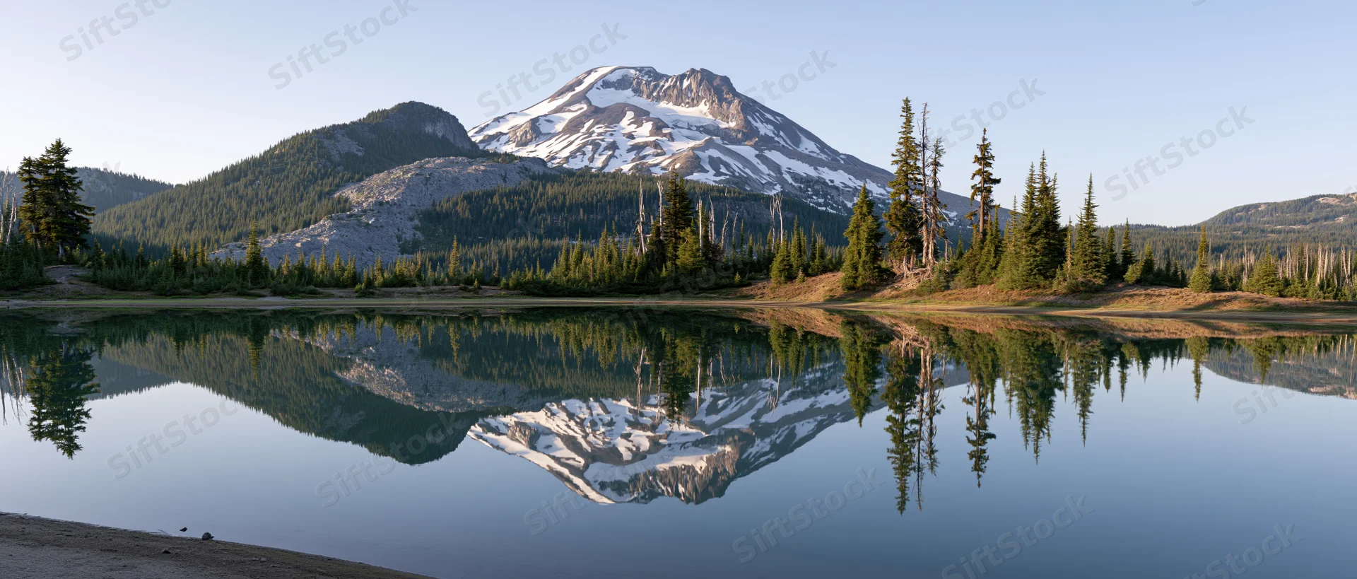 Snow-capped volcanic mountain reflected in calm lake surrounded by evergreen forest
