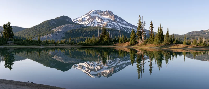 Snow-capped volcanic mountain reflected in calm lake surrounded by evergreen forest