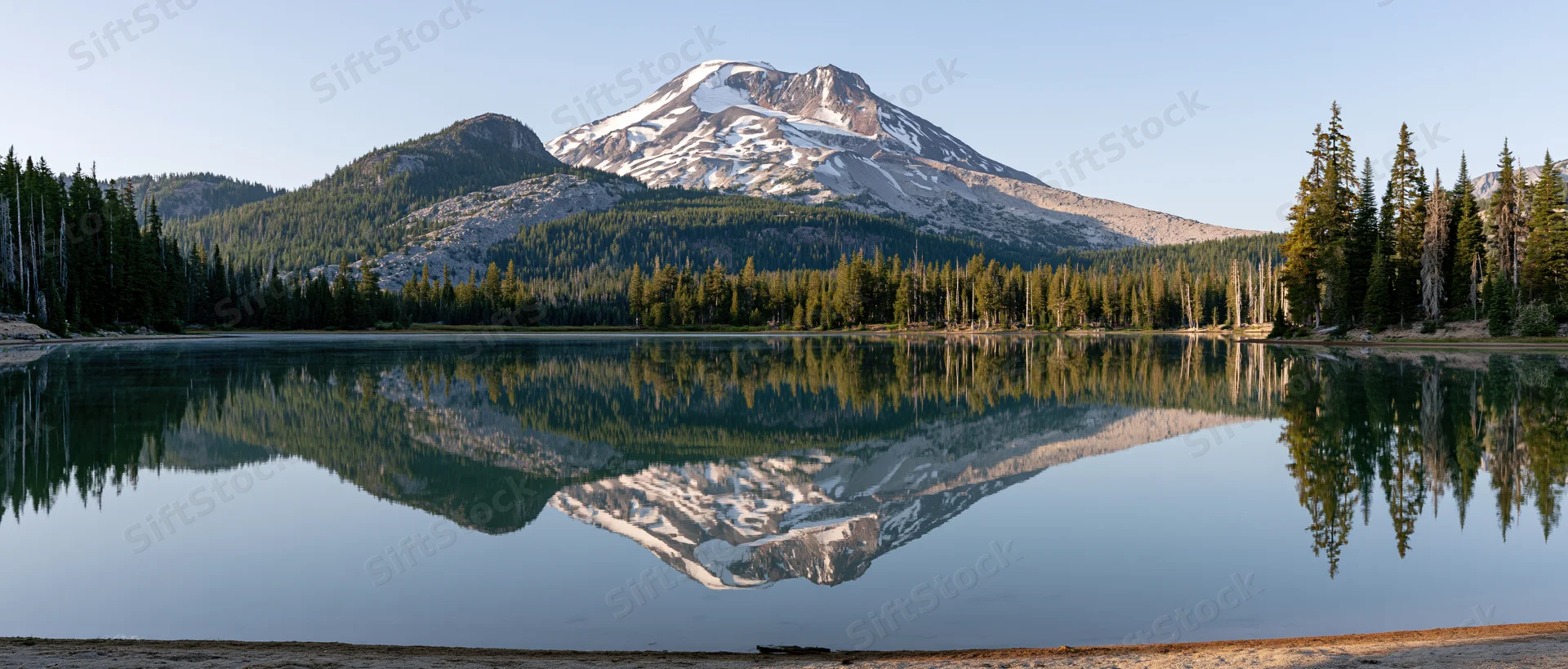 Snow-capped volcanic mountain reflected in calm lake surrounded by pine forest