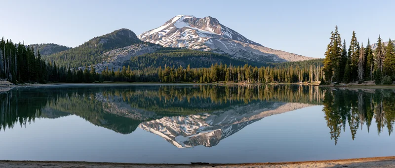 Snow-capped volcanic mountain reflected in calm lake surrounded by pine forest