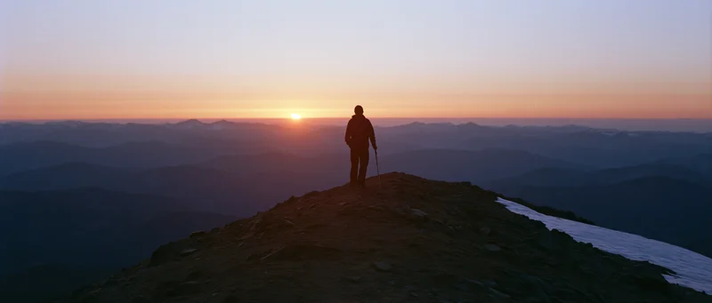 Hiker standing on mountain peak at sunset holding trekking pole