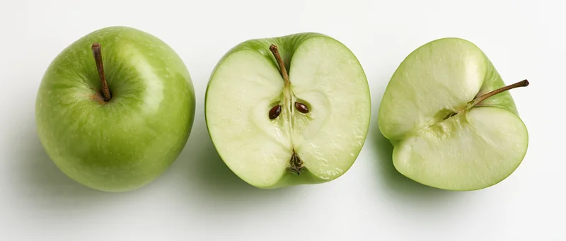 Three green apples in a row, whole, halved, and sliced, on a white surface
