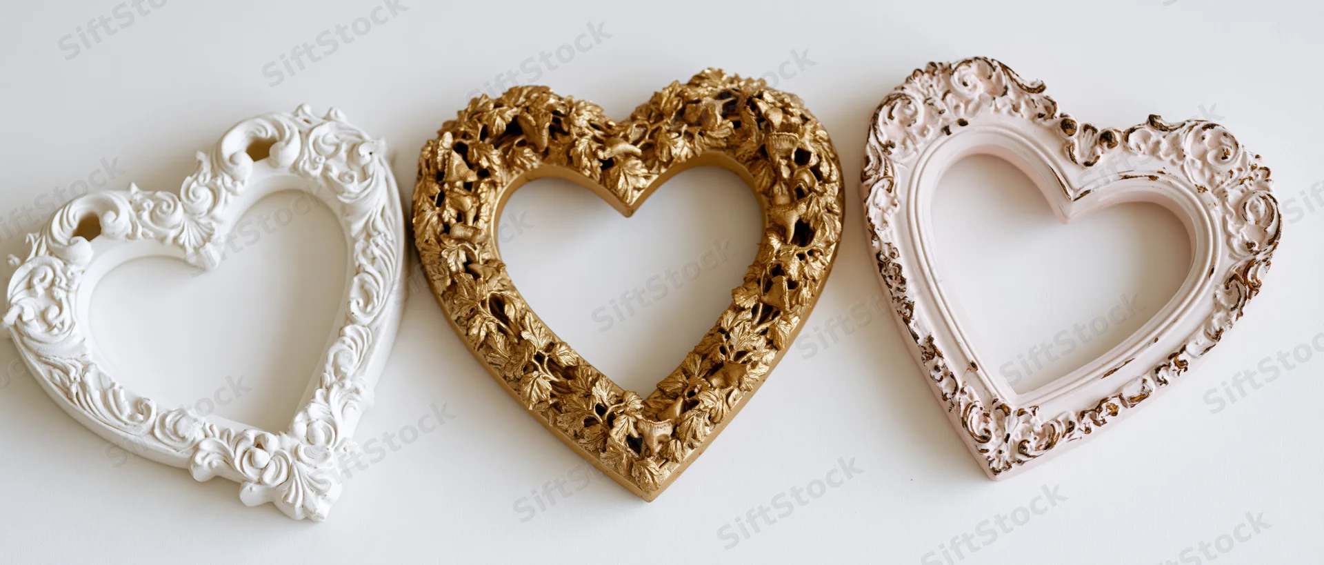 Three ornate baroque heart-shaped frames in white, gold and pink arranged side by side on a white surface