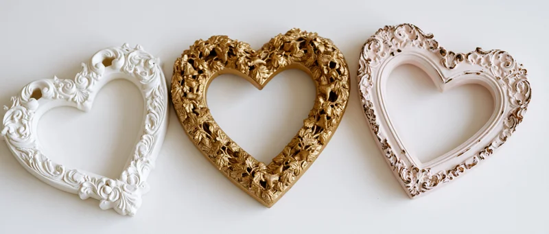 Three ornate baroque heart-shaped frames in white, gold and pink arranged side by side on a white surface
