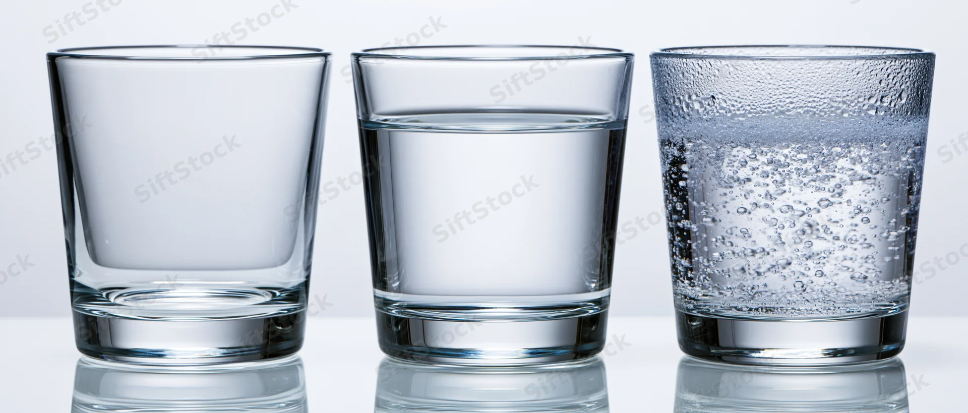 Three short tumbler glasses arranged side by side on a white surface, each containing different states of water from empty to still to carbonated, reflecting light and showing clarity and condensation