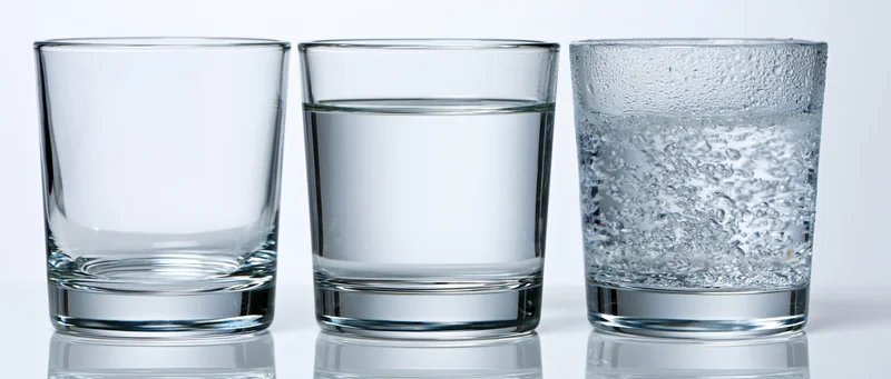 Three short tumbler glasses arranged side by side on a white reflective surface, one empty, one half-full of still water, one full of carbonated water with bubbles and condensation