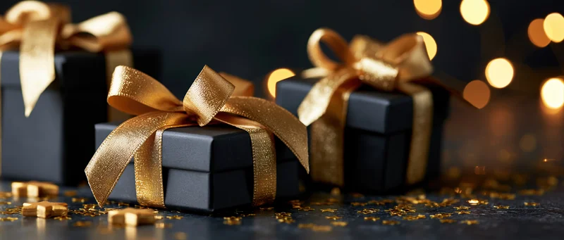 Three black gift boxes with gold satin ribbons arranged on a dark surface with festive lights and glitter in the background