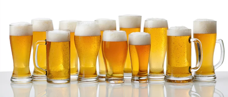 Twelve beer glasses and mugs lined up in a row on a reflective white surface