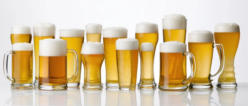 Twelve different beer glasses and mugs lined up in a row each filled with golden beer and white foam on a white reflective surface