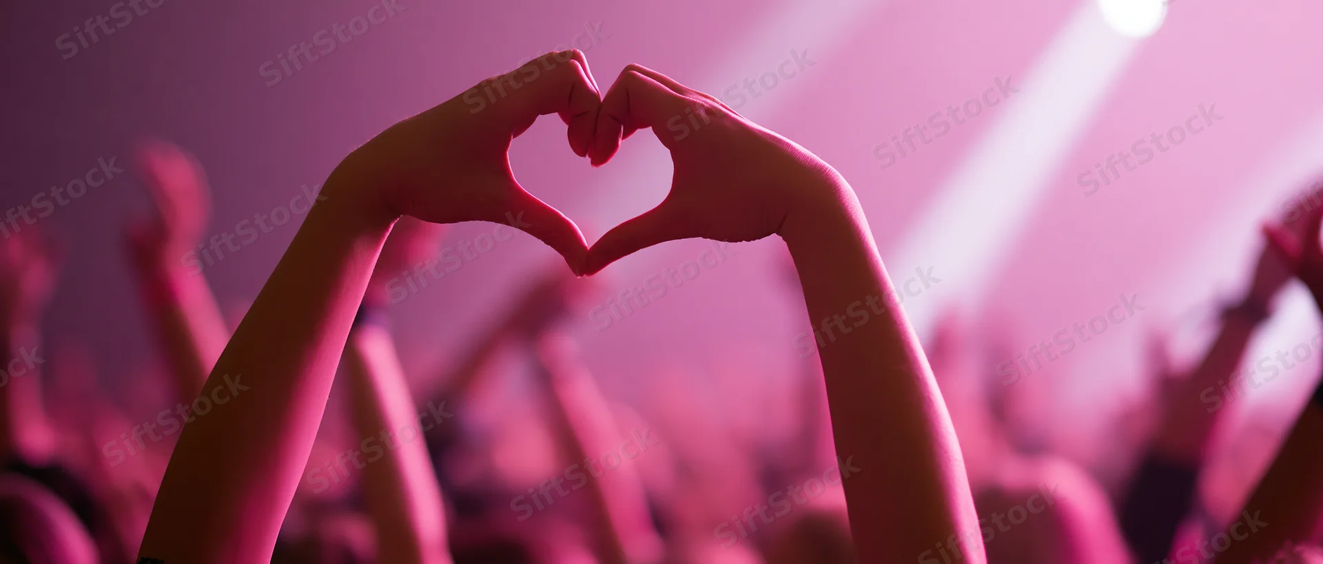 Hands forming heart shape above crowd at concert under pink stage lights