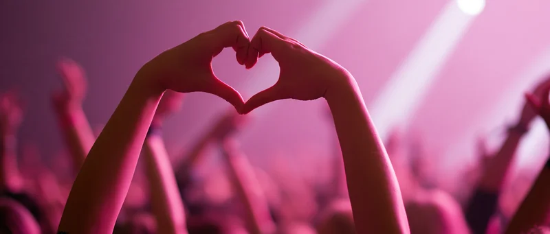 Hands forming heart shape above crowd at concert under pink stage lights