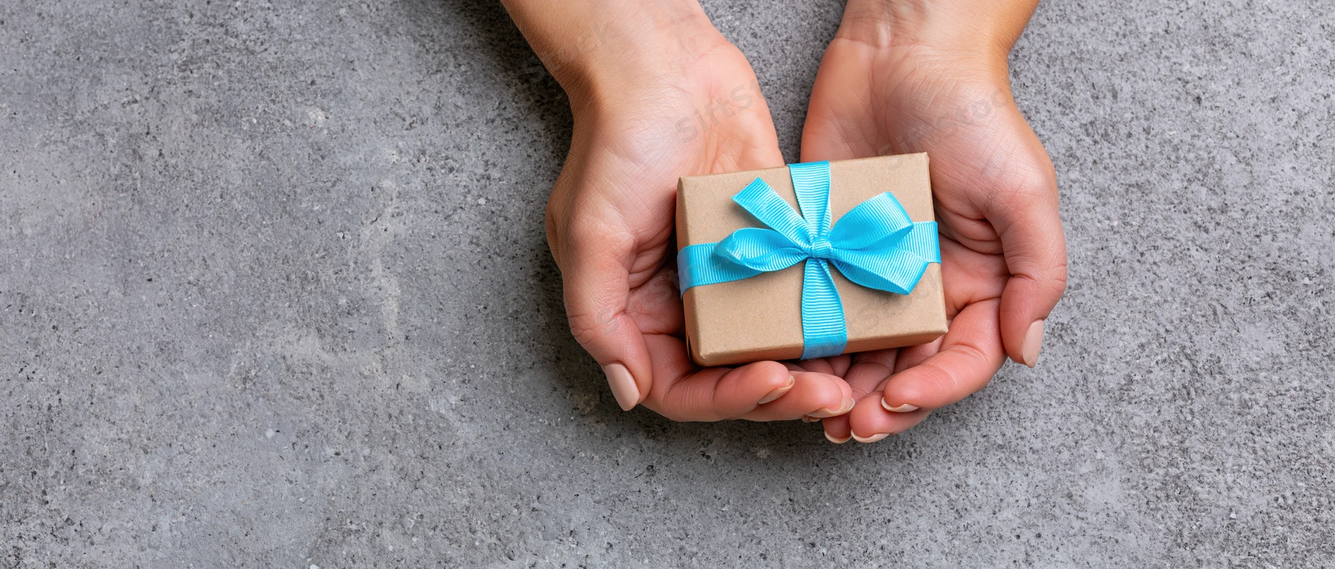 Hands holding a small kraft paper gift box with blue ribbon on gray surface
