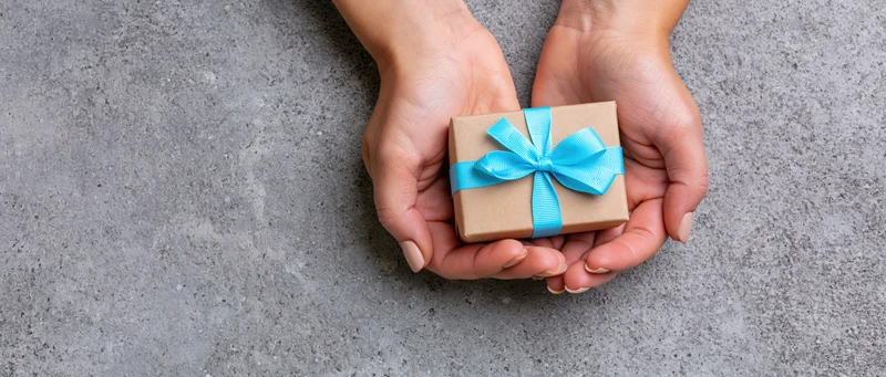 Hands holding a small kraft paper gift box with blue ribbon on gray surface