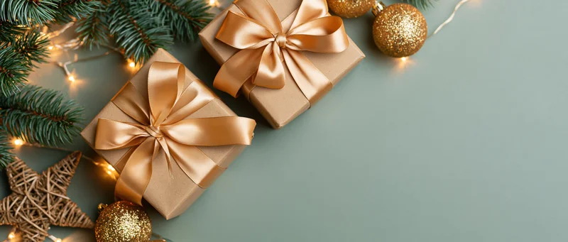 Two kraft paper gift boxes tied with satin champagne ribbons on a flat surface with pine branches, gold ornaments, and string lights, arranged diagonally with copyspace on the right
