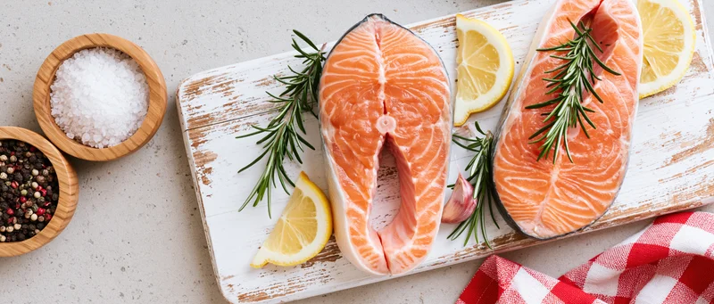 Two raw salmon steaks on a weathered white wooden board with lemon, rosemary, garlic, salt and pepper bowls, red checkered cloth nearby