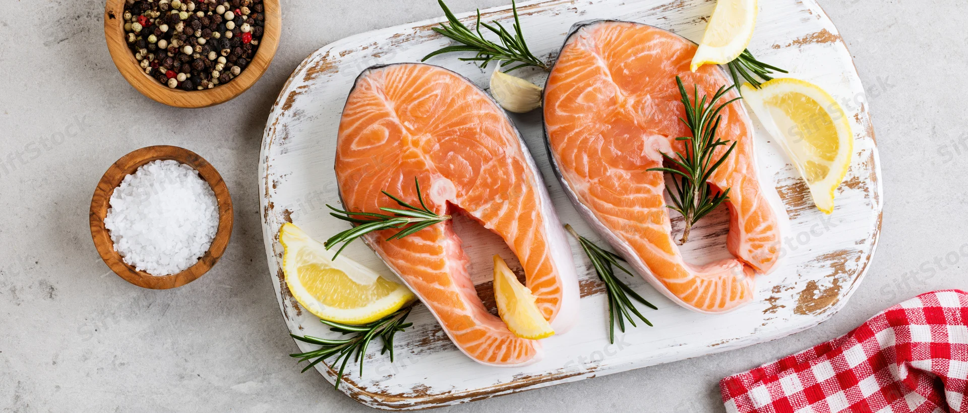 Two raw salmon steaks on a weathered white wooden board with lemon, rosemary, and garlic, surrounded by salt and pepper bowls on a gray countertop
