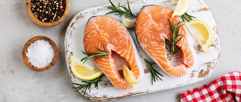 Two raw salmon steaks on a weathered white wooden board with lemon, rosemary, and garlic, surrounded by salt and pepper bowls on a gray countertop