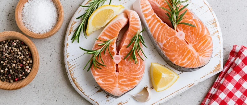 Two raw salmon steaks on a weathered white wooden board with lemon, rosemary, garlic, salt, pepper, and a red checkered napkin on a gray countertop