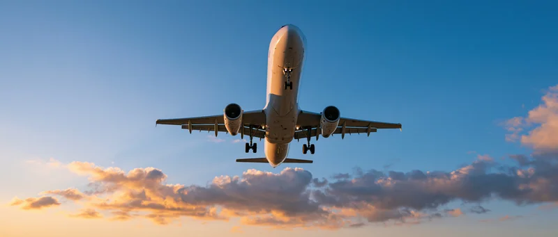 White commercial passenger jet flying overhead with landing gear down against sunset sky