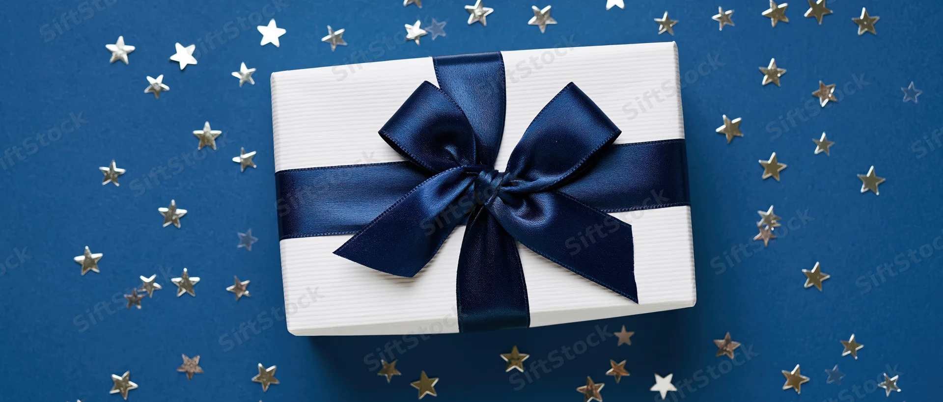White gift box with navy satin ribbon and bow on blue background with scattered stars