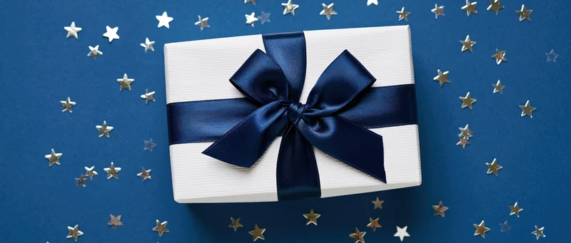 White gift box with navy satin ribbon and bow on blue background with scattered stars