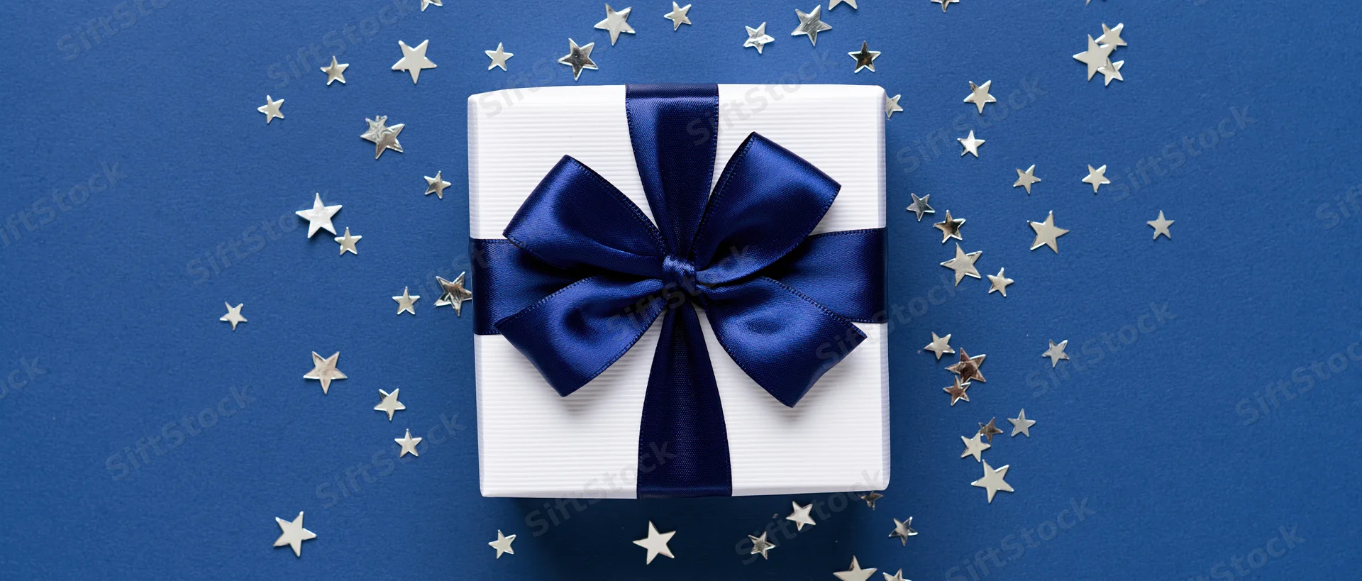 White gift box with navy satin ribbon and bow on blue surface surrounded by silver stars