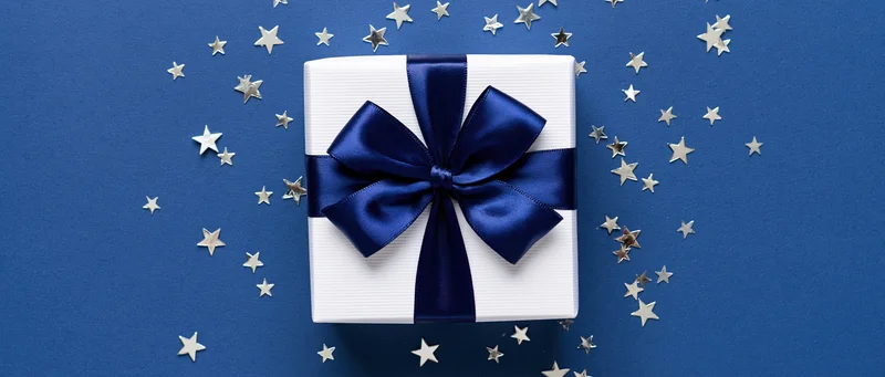 White gift box with navy satin ribbon and bow on blue surface surrounded by silver stars