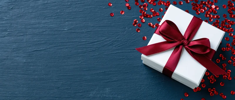 White gift box with red satin ribbon and bow on dark blue textured surface surrounded by red sequins