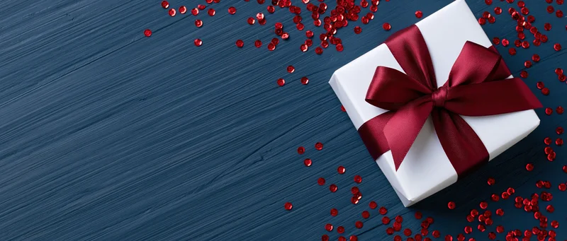 White gift box with red satin ribbon and bow on dark blue wooden surface surrounded by red confetti