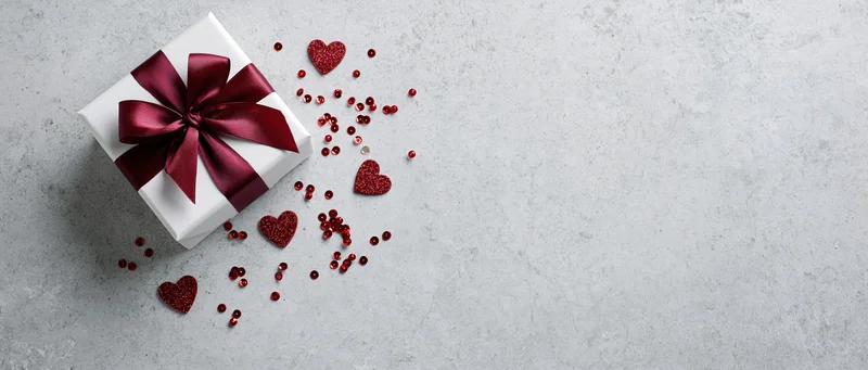White gift box with red satin ribbon and scattered heart-shaped glitter on gray surface