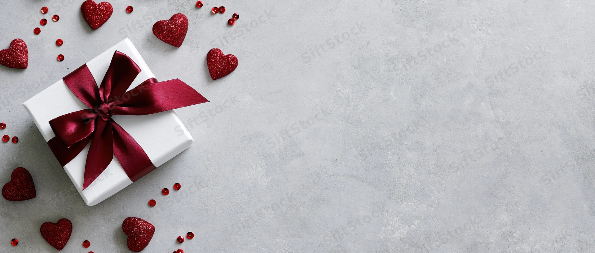 White gift box with red satin ribbon bow and glitter hearts on gray surface