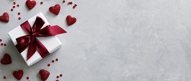 White gift box with red satin ribbon bow and glitter hearts on gray surface
