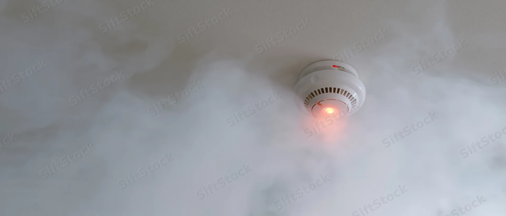 White smoke detector with red light activated on ceiling surrounded by smoke