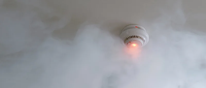 White smoke detector with red light activated on ceiling surrounded by smoke