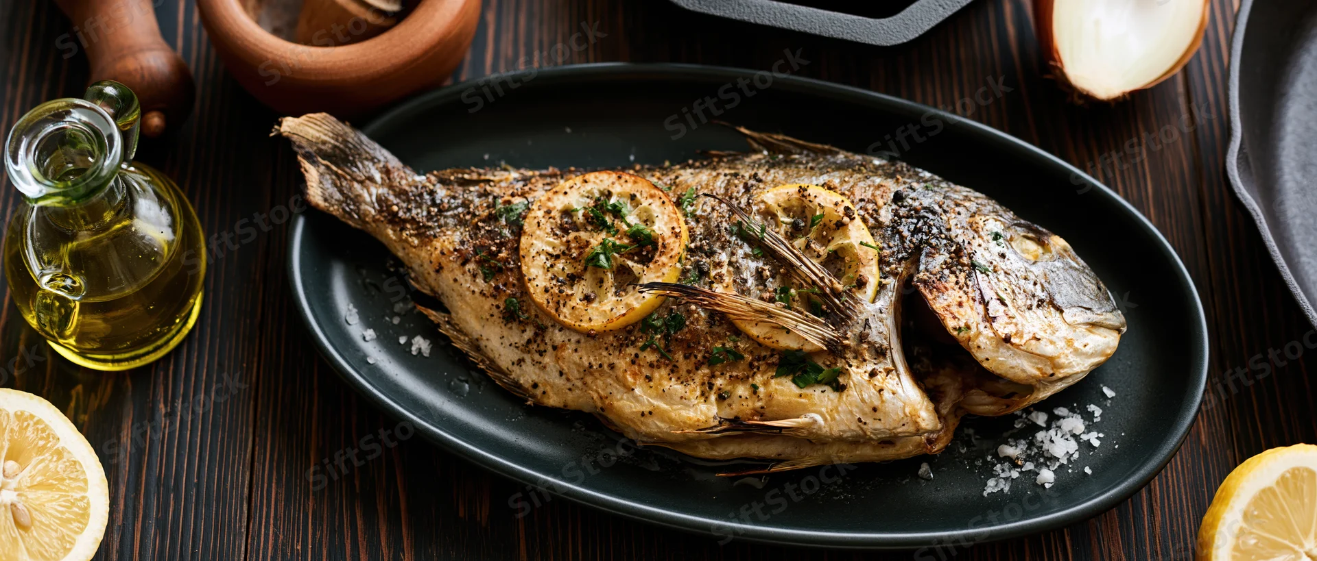 Whole grilled sea bream on dark oval plate with lemon slices and herbs, surrounded by olive oil and salt on wooden table