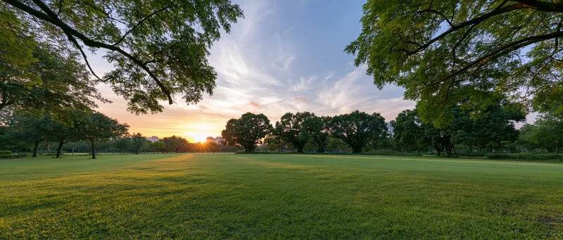Wide panoramic view of a manicured urban park at sunset with green lawn and trees framing the horizon