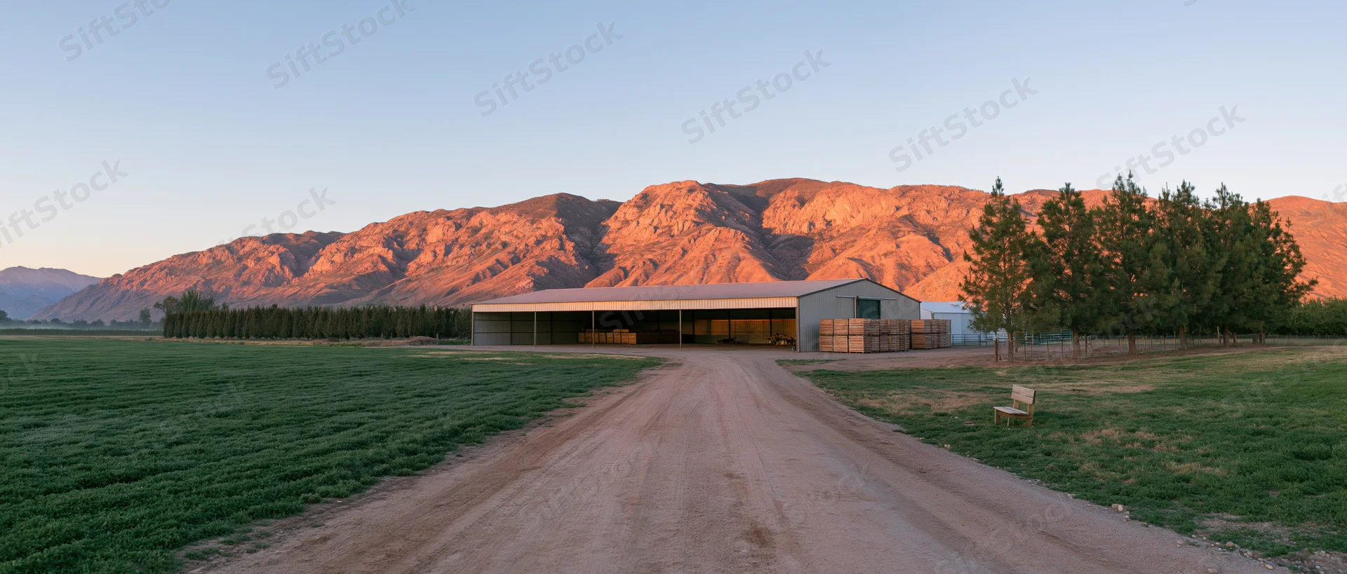 Wide panoramic view of a rural farm with dirt road leading to a barn at golden hour against mountains