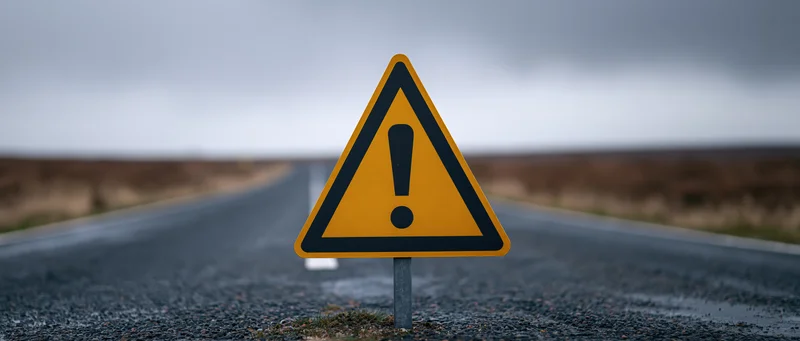 Yellow triangular warning sign with exclamation mark on roadside of empty road