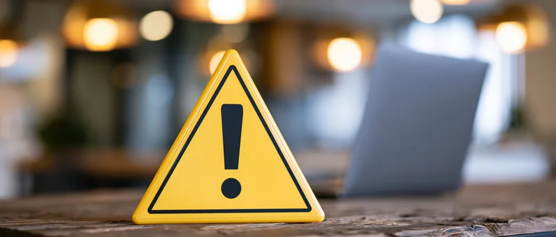 Yellow triangular warning sign with exclamation mark on wooden desk near laptop in office setting