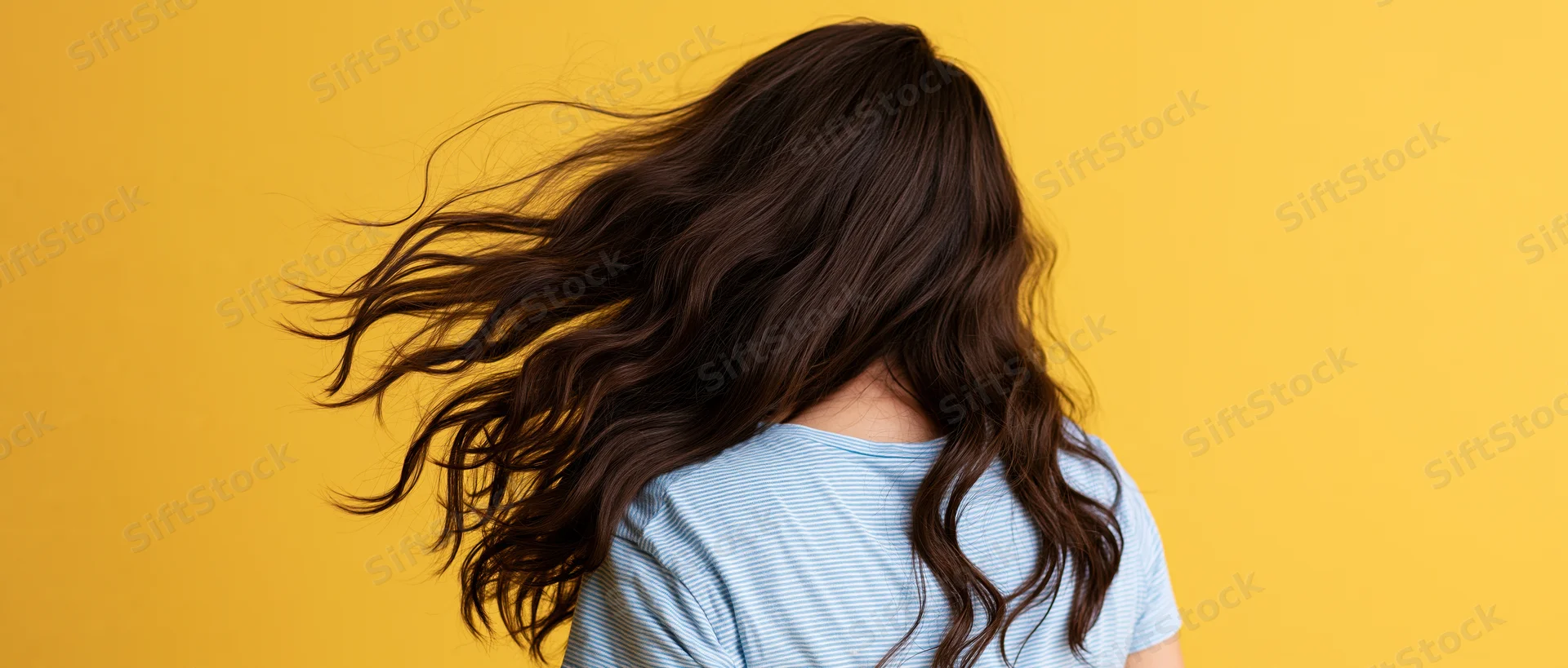 Woman seen from back with long dark brown wavy hair flowing in motion against yellow background