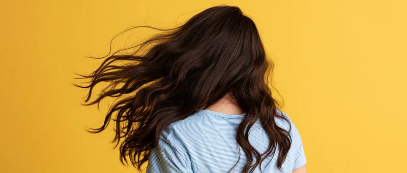 Woman seen from back with long dark brown wavy hair flowing in motion against yellow background
