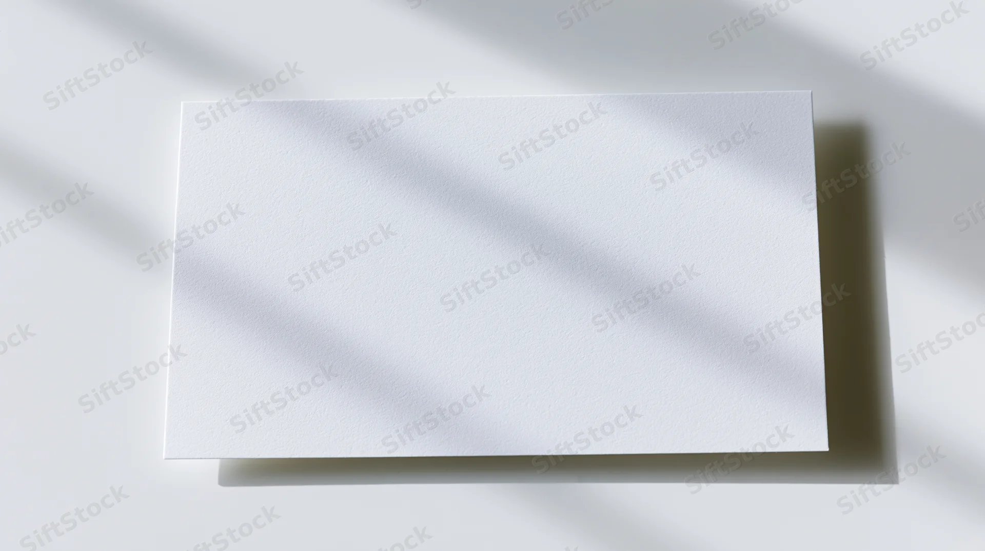 Blank white card lying flat on a white surface with diagonal shadows, top view