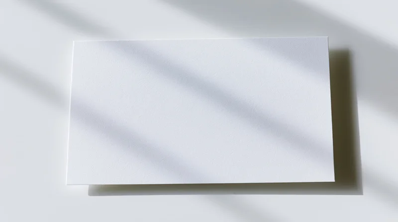 Blank white card lying flat on a white surface with diagonal shadows, top view