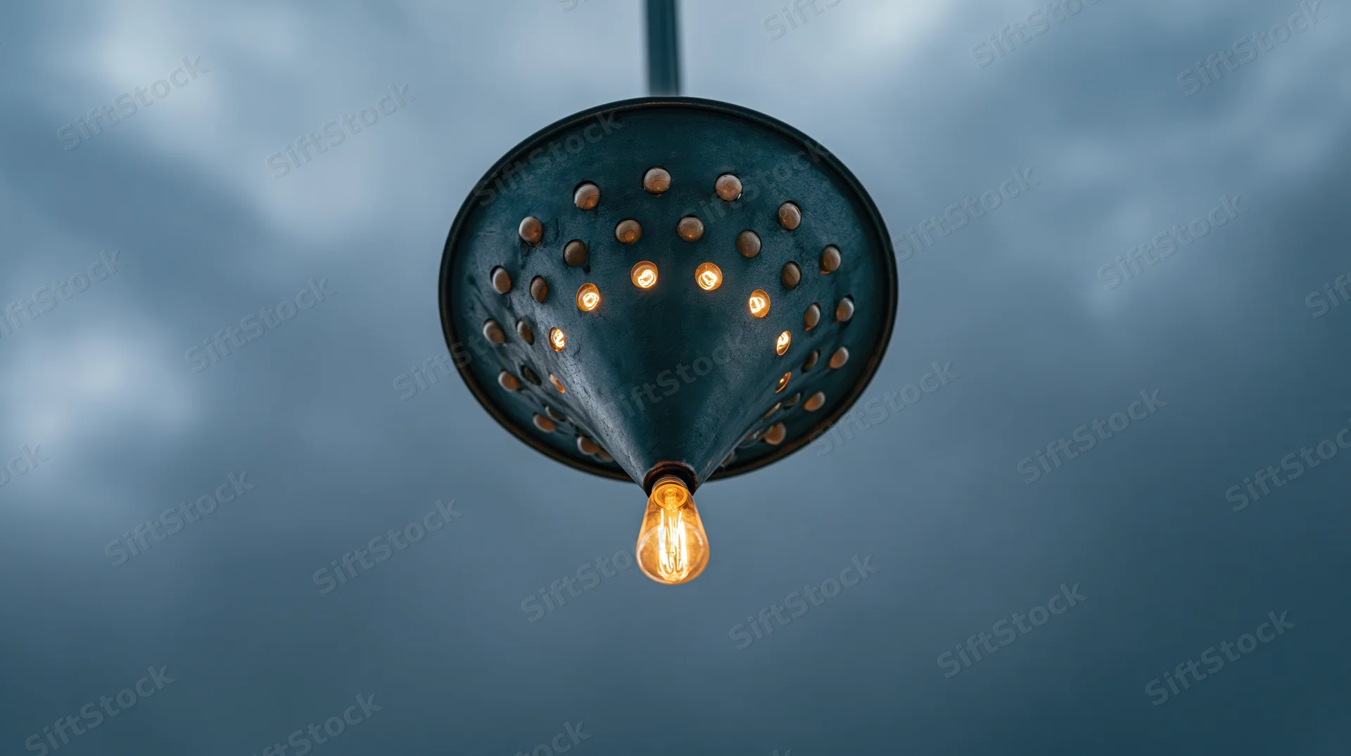Dark perforated metal funnel with glowing edison bulb hanging against cloudy sky