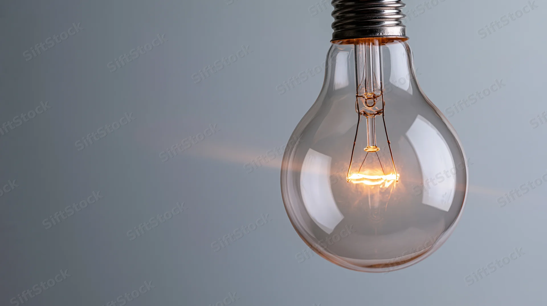 Glowing incandescent lightbulb hanging against a plain gray background with visible tungsten filament and warm light emission