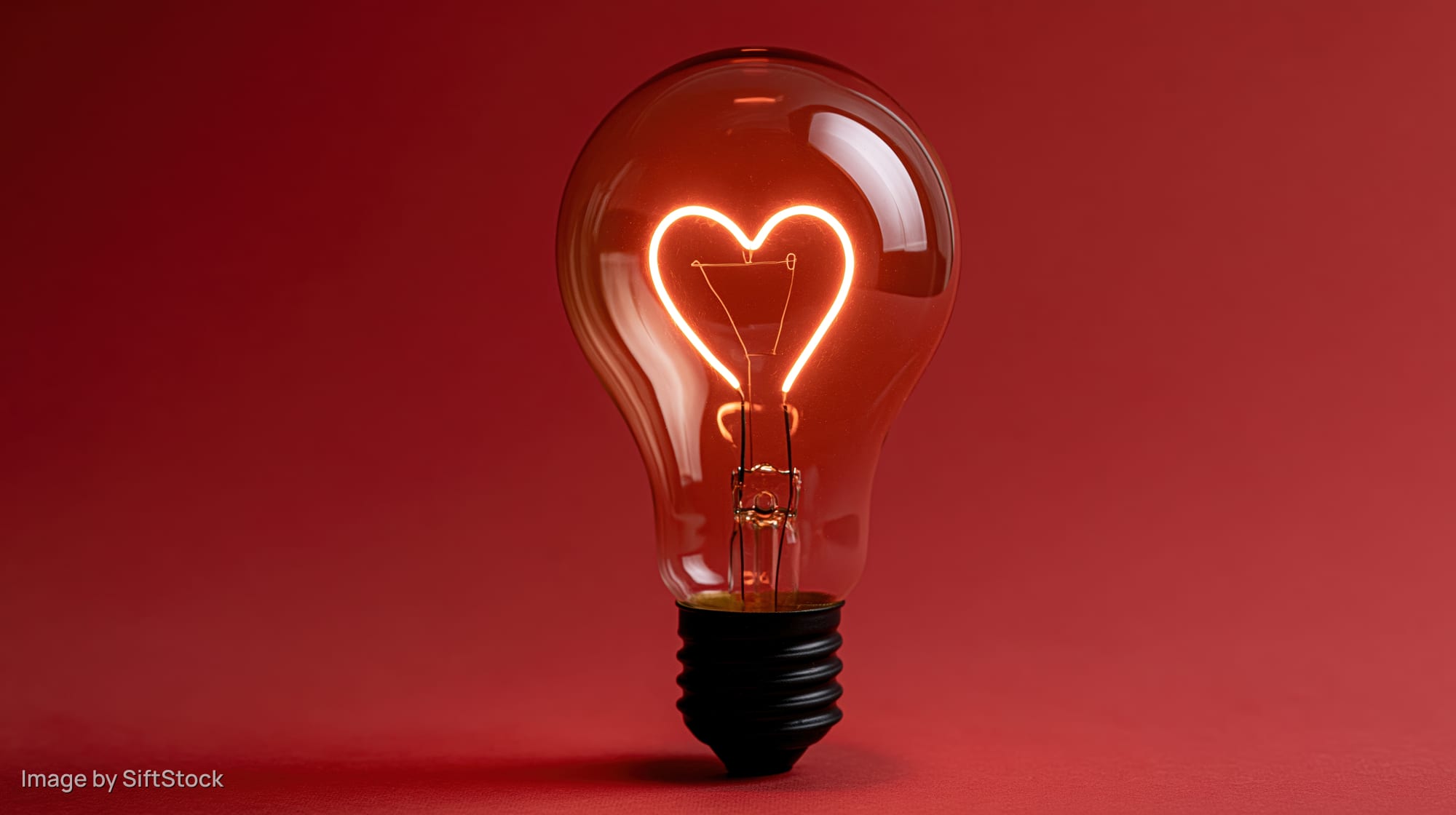 Glowing heart-shaped filament inside a glass lightbulb on red background