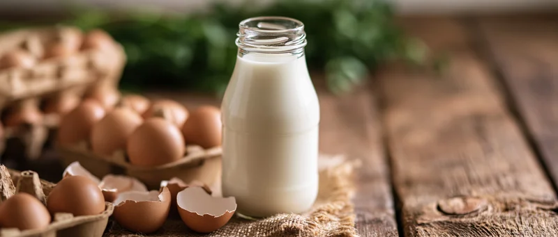 Glass milk bottle filled with fresh milk on wooden table beside eggs and eggshells